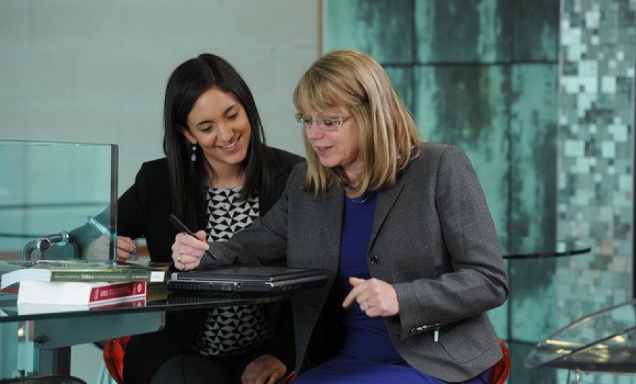 A professor sits at a glass table holding a pen, and there is a student beside her listening to her.